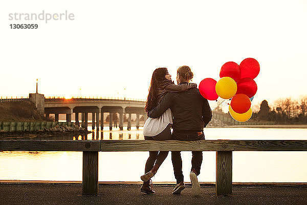 Rückansicht eines Paares mit Luftballons  das bei Sonnenuntergang auf einem Geländer sitzt