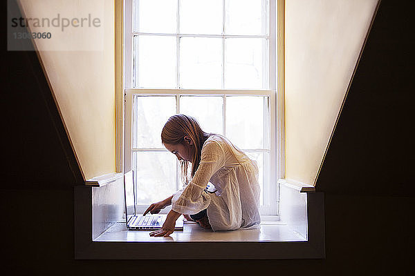 Mädchen benutzt Laptop-Computer  während sie zu Hause am Fenster sitzt