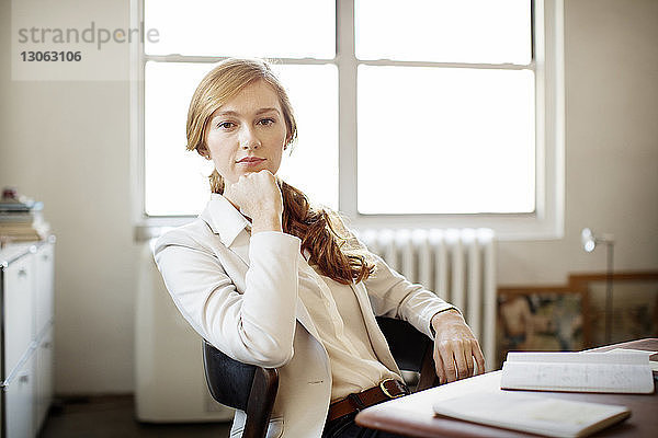 Porträt einer selbstbewussten Geschäftsfrau mit Hand am Kinn am Schreibtisch sitzend im Büro