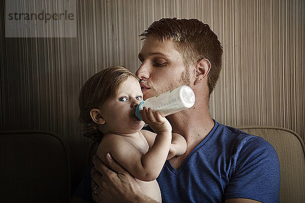 Säugling trinkt Milch  während der Vater ihn zu Hause küsst