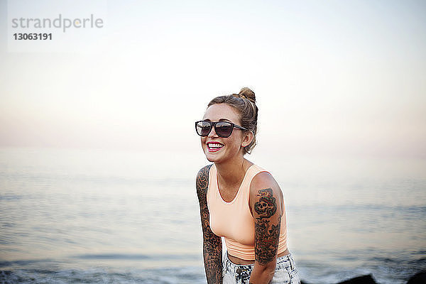 Glückliche Frau mit Sonnenbrille steht am Strand gegen den Himmel