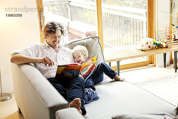 Glückliche Vater und Sohn lesen Buch  während sie zu Hause auf dem Sofa sitzen