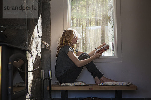 Frau liest Buch  während sie zu Hause am Fenster sitzt