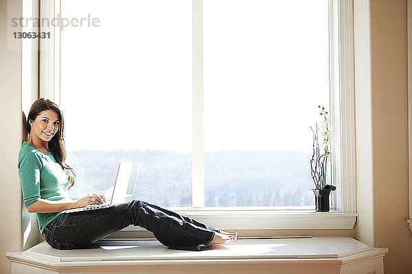 Seitenansicht einer Frau  die am Fenster sitzt und einen Laptop benutzt