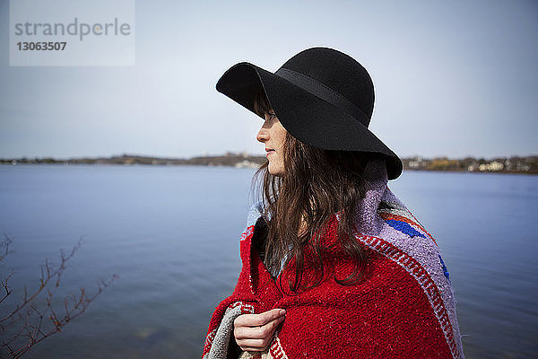 Nachdenkliche Frau mit Hut und Decke am Seeufer stehend