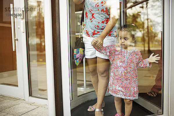 Ausgeschnittenes Bild einer Mutter  die ihre Tochter an der Hand hält  während sie an einer Glastür steht