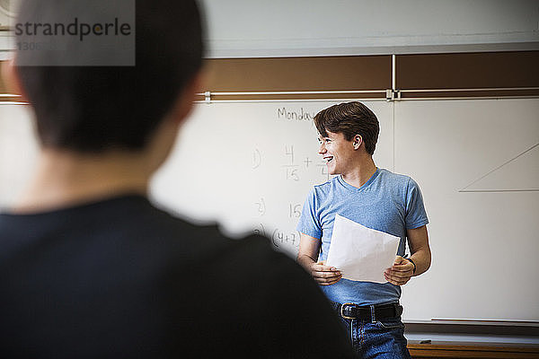 Mann hält Papier  während er im Klassenzimmer gegen die Tafel steht