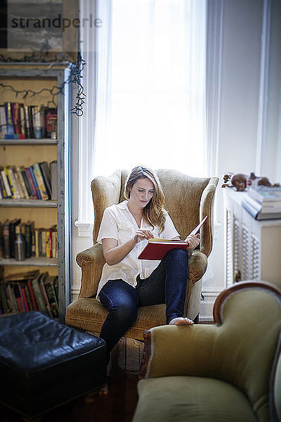 Frau liest Buch  während sie zu Hause auf einem Sessel sitzt