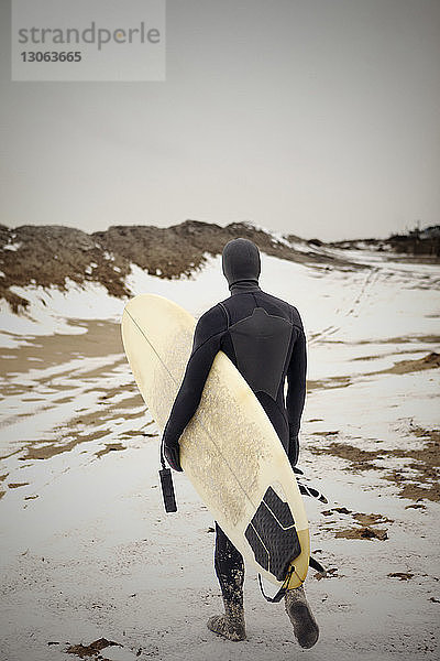Rückansicht eines Mannes  der ein Surfbrett trägt  während er auf einem schneebedeckten Feld geht