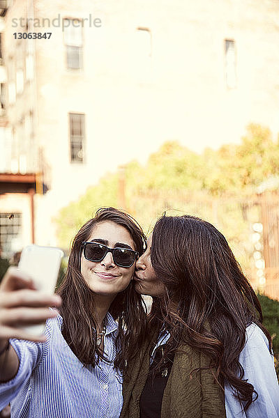 Mutter küsst Tochter  während sie an einem sonnigen Tag Selbstgespräche per Smartphone führt