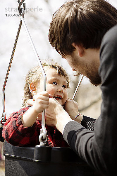 Glücklicher Vater schaut auf Tochter  die auf einer Schaukel auf dem Spielplatz sitzt