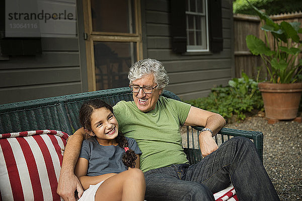 Glücklicher Großvater und glückliche Enkelin sitzen auf einem Korbsofa im Hof