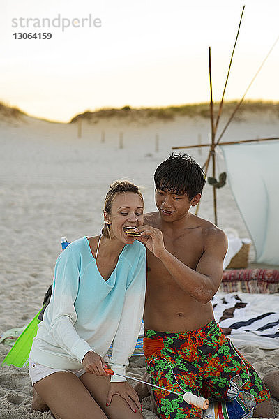 Mann füttert Freundin mit Keksen  während er am Strand am Lagerfeuer kniet