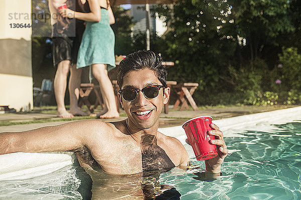 Glücklicher Mann ohne Hemd mit Sonnenbrille  der ein Getränk hält  während er im Pool steht