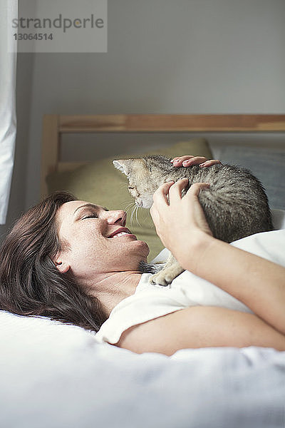 Lächelnde Frau spielt mit Katze  während sie zu Hause auf dem Bett liegt