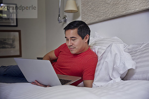 Mann benutzt Laptop-Computer  während er zu Hause auf dem Bett liegt