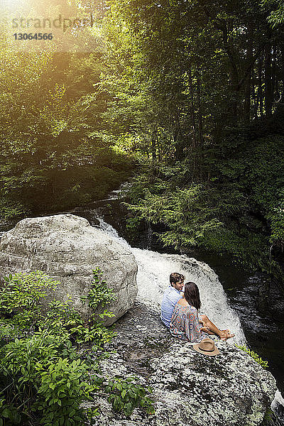 Hochwinkelaufnahme eines Paares  das auf Felsen am Wasserfall im Wald sitzt