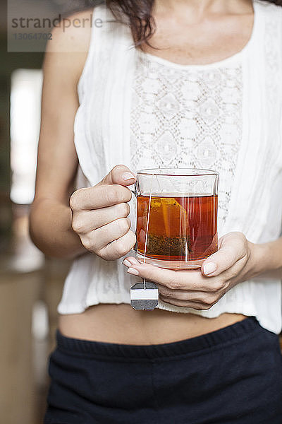 Mittelteil einer Frau  die zu Hause stehend eine Tasse mit Teebeutel hält