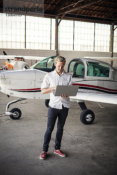 Mann benutzt Laptop-Computer  während er im Hangar an einem Propellerflugzeug steht