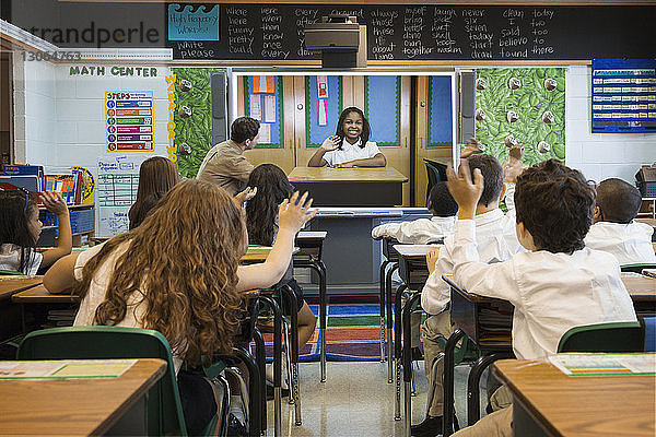 Rückansicht von Schülern  die mit den Händen winken  während sie ein Foto auf einer Leinwand im Klassenzimmer betrachten