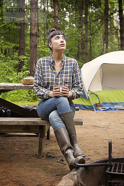 Frau schaut auf  während sie auf einem Picknicktisch vor einem Zelt im Wald sitzt
