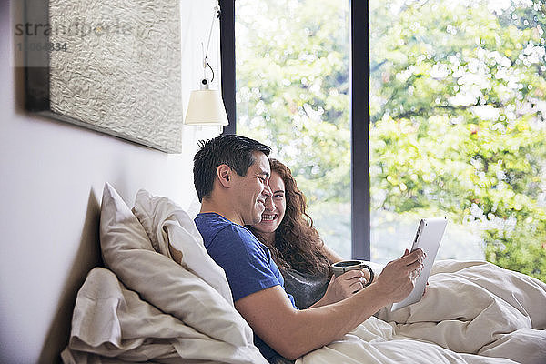 Glückliches Paar  das sich unterhält und einen Tablet-Computer benutzt  während es zu Hause im Bett liegt