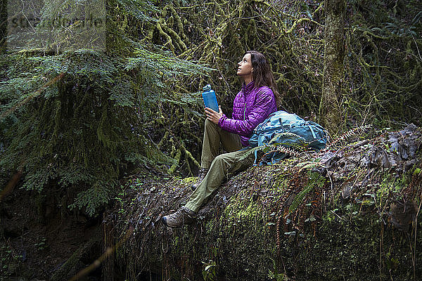 Frau hält Flasche und schaut auf  während sie im Wald auf einem umgefallenen Baumstamm sitzt