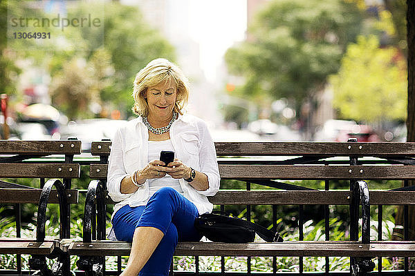 Glückliche Frau benutzt Mobiltelefon  während sie in der Stadt auf einer Bank sitzt