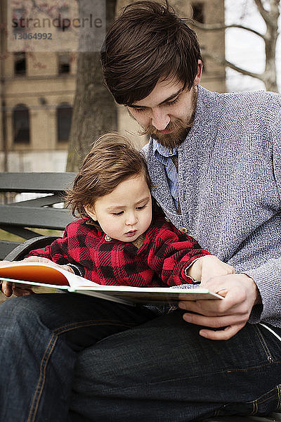 Mann liest Buch  während er mit seiner Tochter auf einer Parkbank sitzt