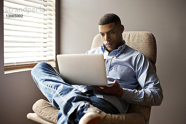 Mann benutzt Laptop-Computer  während er zu Hause auf einem Stuhl sitzt