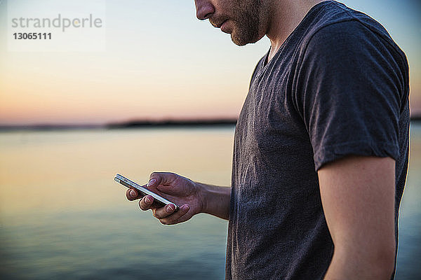 Mittelsektion eines Mannes  der während des Sonnenuntergangs am Meer ein Mobiltelefon benutzt