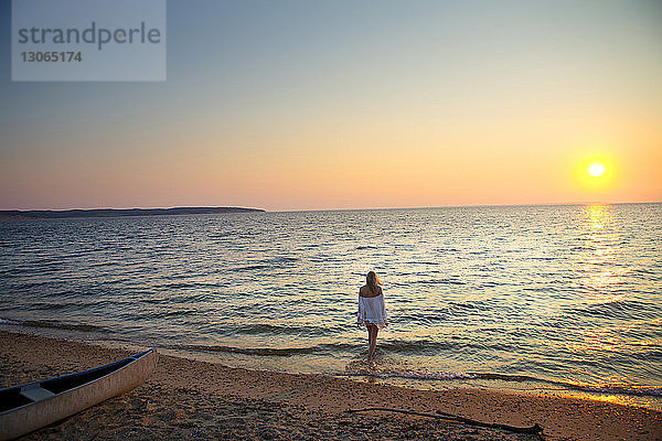 Rückansicht einer Frau  die bei Sonnenuntergang am Strand am Ufer spazieren geht