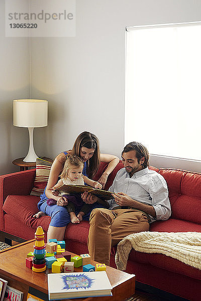 Vater und Mutter zeigen einem Mädchen ein Bilderbuch  während sie zu Hause auf dem Sofa sitzen