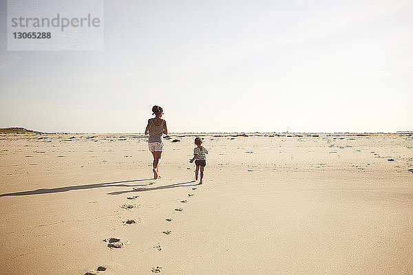 Mutter und Tochter rennen am Strand gegen den klaren Himmel