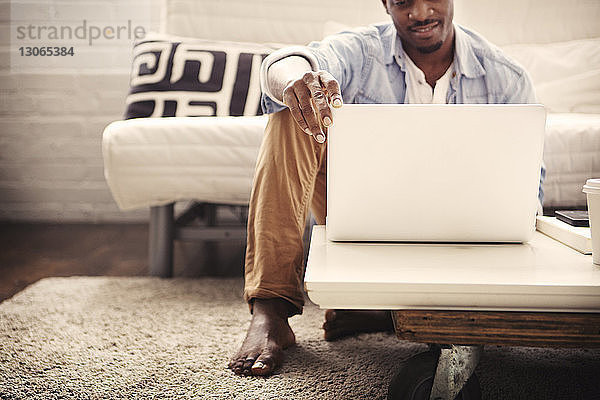 Lächelnder Mann benutzt Laptop-Computer  während er zu Hause auf dem Teppich sitzt