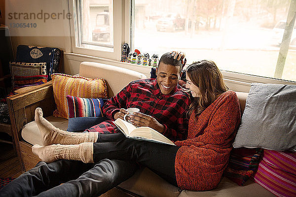 Ein Paar liest ein Buch  während es zu Hause auf dem Sofa Kaffee trinkt