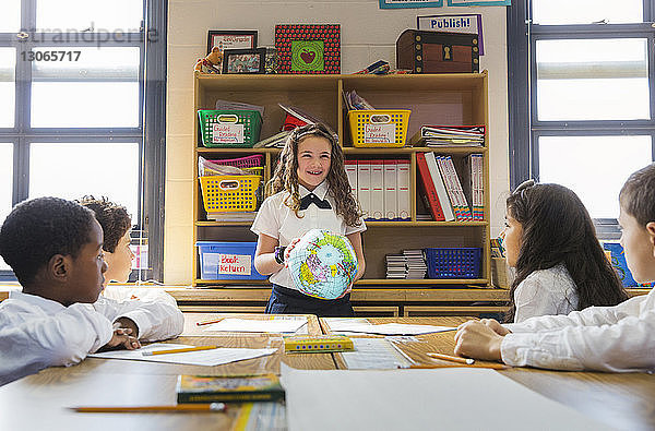 Schülerinnen betrachten ein Mädchen  das während des Unterrichts im Klassenzimmer einen Globus hält