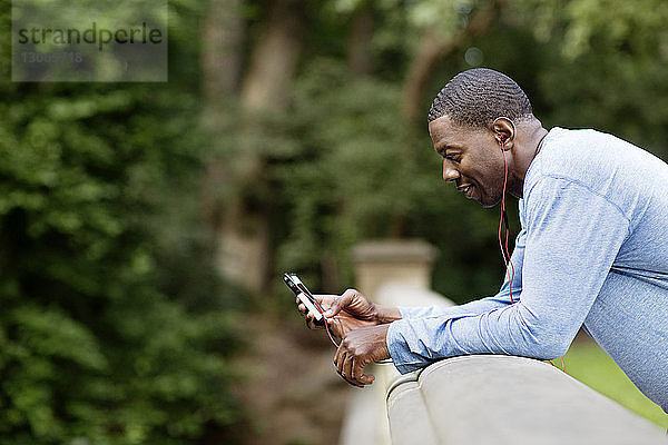 Seitenansicht eines Mannes  der ein Smartphone benutzt  während er sich an eine Stützmauer im Park lehnt