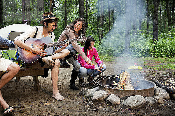 Frau röstet Marshmallows  während sie am Lagerfeuer im Wald bei einem Freund sitzt und Gitarre spielt