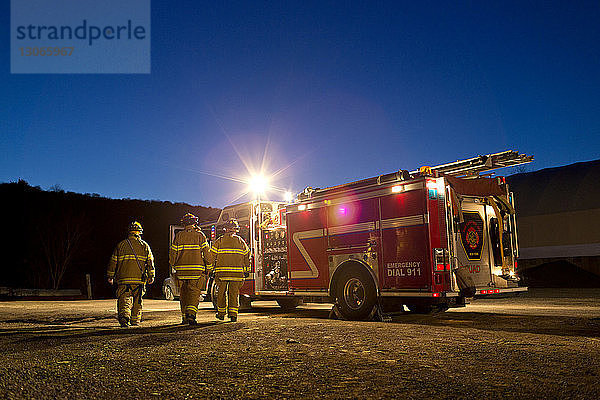 Rückansicht der Feuerwehrleute  die nachts beim Löschfahrzeug stehen