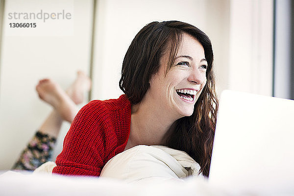Fröhliche Frau mit Laptop-Computer  die zu Hause auf dem Bett liegt