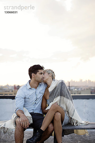 Romantisches  in eine Decke gehülltes Paar ruht auf Gebäudeterrasse gegen den Himmel