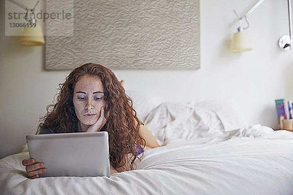 Frau schaut auf Tablet-Computer  während sie im Bett an der Wand liegt