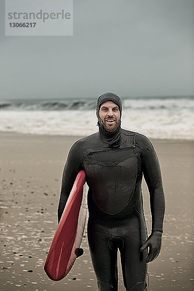 Porträt eines glücklichen Mannes  der ein Surfbrett trägt  während er am Strand steht