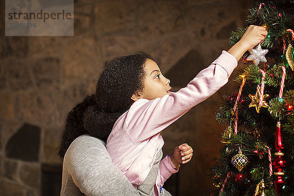 Seitenansicht einer Mutter  die eine Tochter trägt  die zu Hause den Weihnachtsbaum schmückt