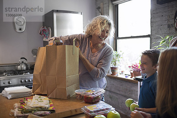 Aufgeregte Kinder sehen Großmutter an  wie sie in der Küche Lebensmittel aus der Einkaufstasche nimmt