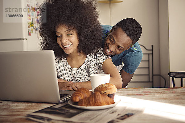 Romantisches Paar sitzt am Tisch mit Croissants und Laptop-Computer in der Küche