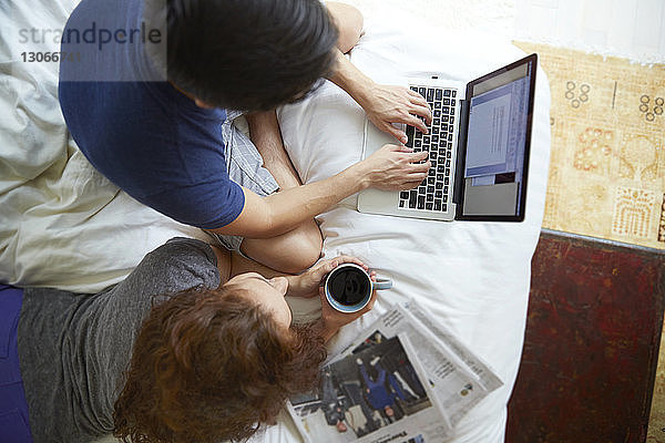 Frau hält Kaffeetasse in der Hand  während ein Mann im Bett liegt und seinen Laptop auf dem Bett benutzt