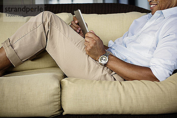 Mittelsektion eines Mannes  der einen Tablet-Computer benutzt  während er sich auf dem Sofa in der Veranda entspannt