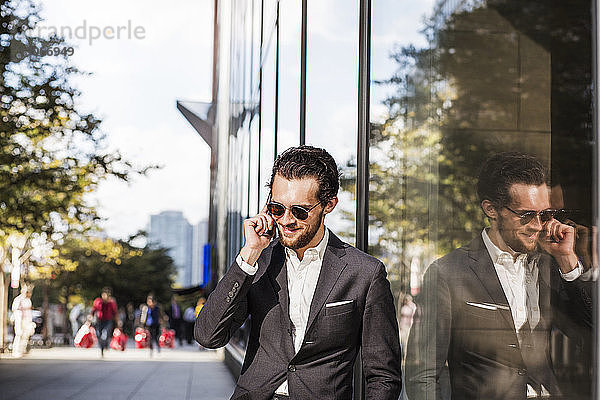 Lächelnder Geschäftsmann benutzt Telefon  das auf Glasgebäude in der Stadt reflektiert